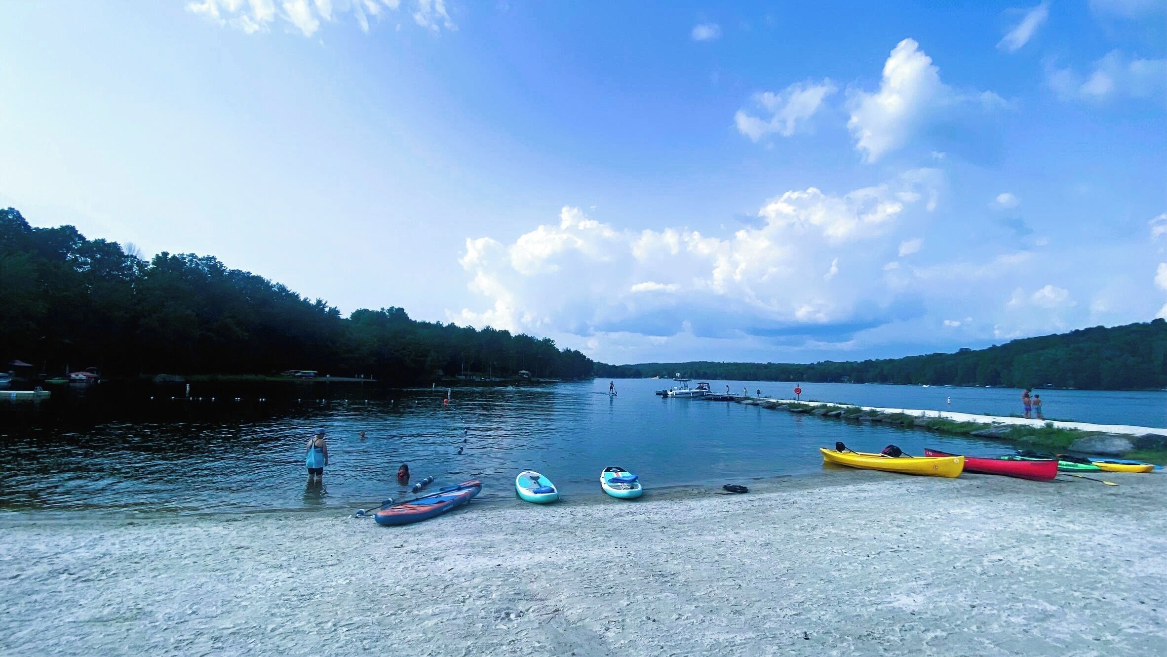 Une scène au bord du lac, avec des kayaks et une planche à pagaie sur la rive, un quai avec des bateaux, et une colline boisée en arrière-plan.
