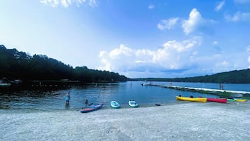 Une scène au bord du lac, avec des kayaks et une planche à pagaie sur la rive, un quai avec des bateaux, et une colline boisée en arrière-plan.