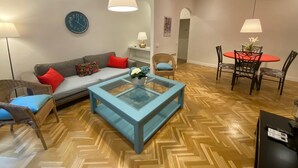 Una sala de estar con sofá, mesa de centro y sillas. Se observa una mesa de comedor con sillas y un reloj en la pared.