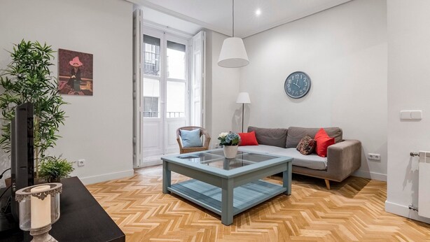 Una sala de estar moderna con una mesa de centro de vidrio, un sofá con cojines rojos y azules, un sillón de mimbre y una planta grande.