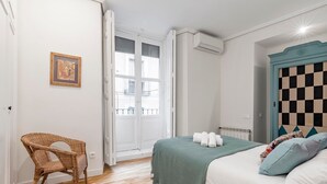 Un dormitorio con cama, una silla de mimbre, cabecera a cuadros y una ventana con persianas.