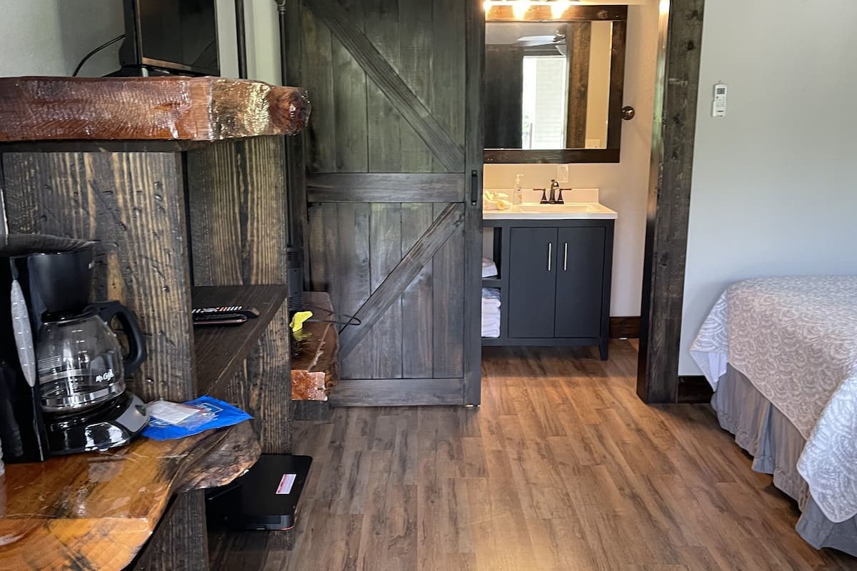 A room with a wooden floor, a bed, a desk with a coffee maker, a mirror, and a sliding door leading to a bathroom.