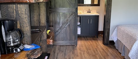 Ein Zimmer mit Holzboden, einem Bett, einem Schreibtisch mit Kaffeemaschine, einem Spiegel und einer Schiebetür zum Badezimmer.