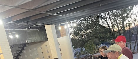 Un grupo de personas cenando en un restaurante al aire libre, con una mesa y taburetes de concreto, rodeado por un muro de ladrillo y árboles.