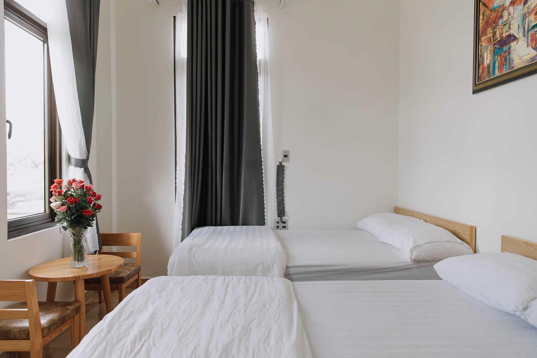 A hotel room with two beds, a wooden table with chairs, a vase of flowers, and a painting on the wall.