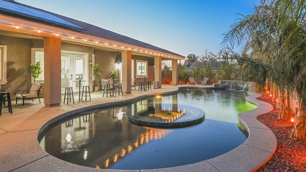 A pool area with a curved design, a covered seating area with chairs and tables, and a building with a flat roof.