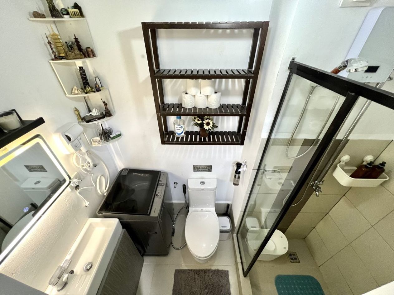 Un baño pequeño con inodoro, lavamanos y zona de ducha. Hay repisas sobre el inodoro con artículos de aseo y objetos decorativos.