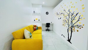 Un salon moderne avec un canapé jaune, un bureau et une fresque murale représentant un arbre dont les feuilles tombent.