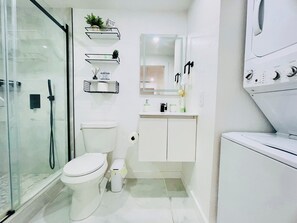 A modern bathroom with a glass shower enclosure, a toilet, a sink with a mirror, and a washing machine. - Magnolias Suite (Lehigh Acres)