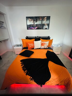 A bed with an orange and black bedspread, two white pillows, and a black throw blanket. There is a white nightstand with a lamp and a box on it. Above the bed, there is a wall-mounted shelf with a few items on it. - Magnolias Suite (Lehigh Acres)