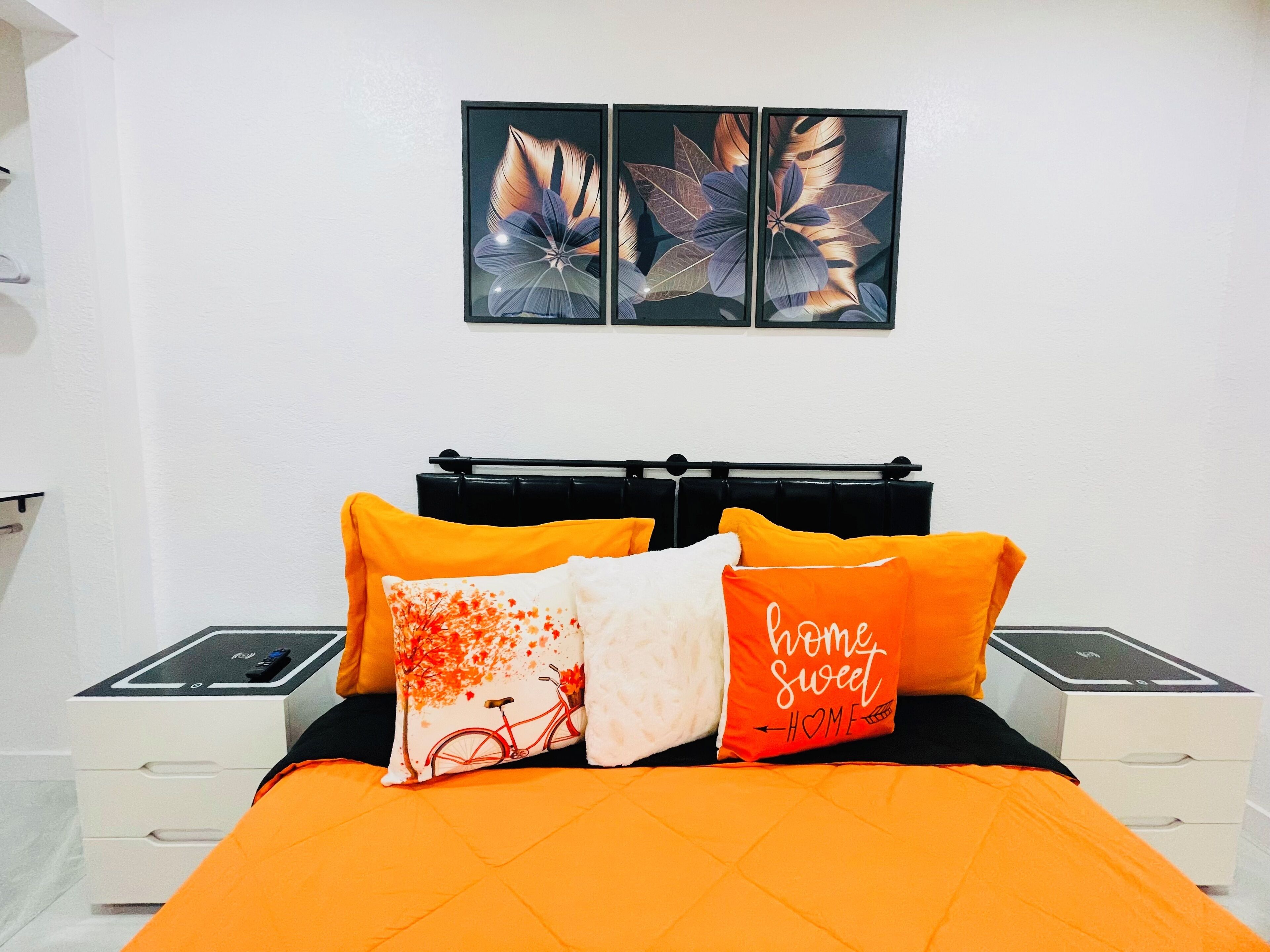 A bed with an orange quilt, black headboard, and four pillows with various designs. There are two bedside tables with electronic devices. Above the bed, there are two framed floral artworks on the wall.