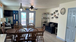 A dining area with a round table and wooden chairs, a ceiling fan, and a kitchen area with a TV and cabinets. - Villa Cape San Blas - Gulf View Townhome, Steps from the Beach & Local Dining (cape san blaste)