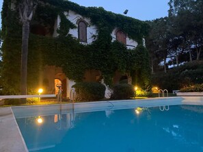 A swimming pool with a stone wall and arched windows, surrounded by greenery and evening lighting. - Villa Loredana in Fontane Bianche (Syracuse)
