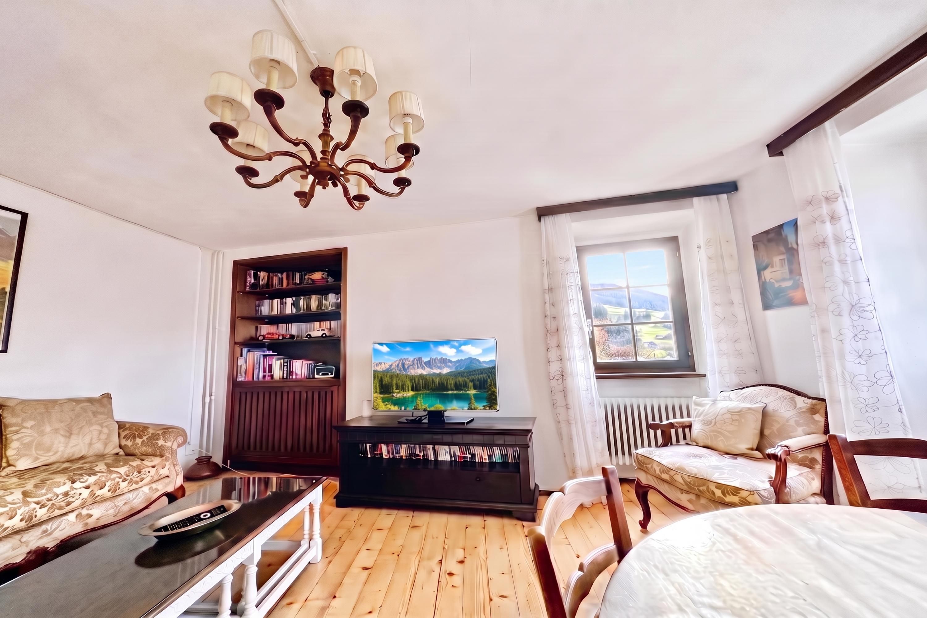 Een woonkamer met houten vloer, een bank, een salontafel, een televisie, een boekenkast en een raam met uitzicht.