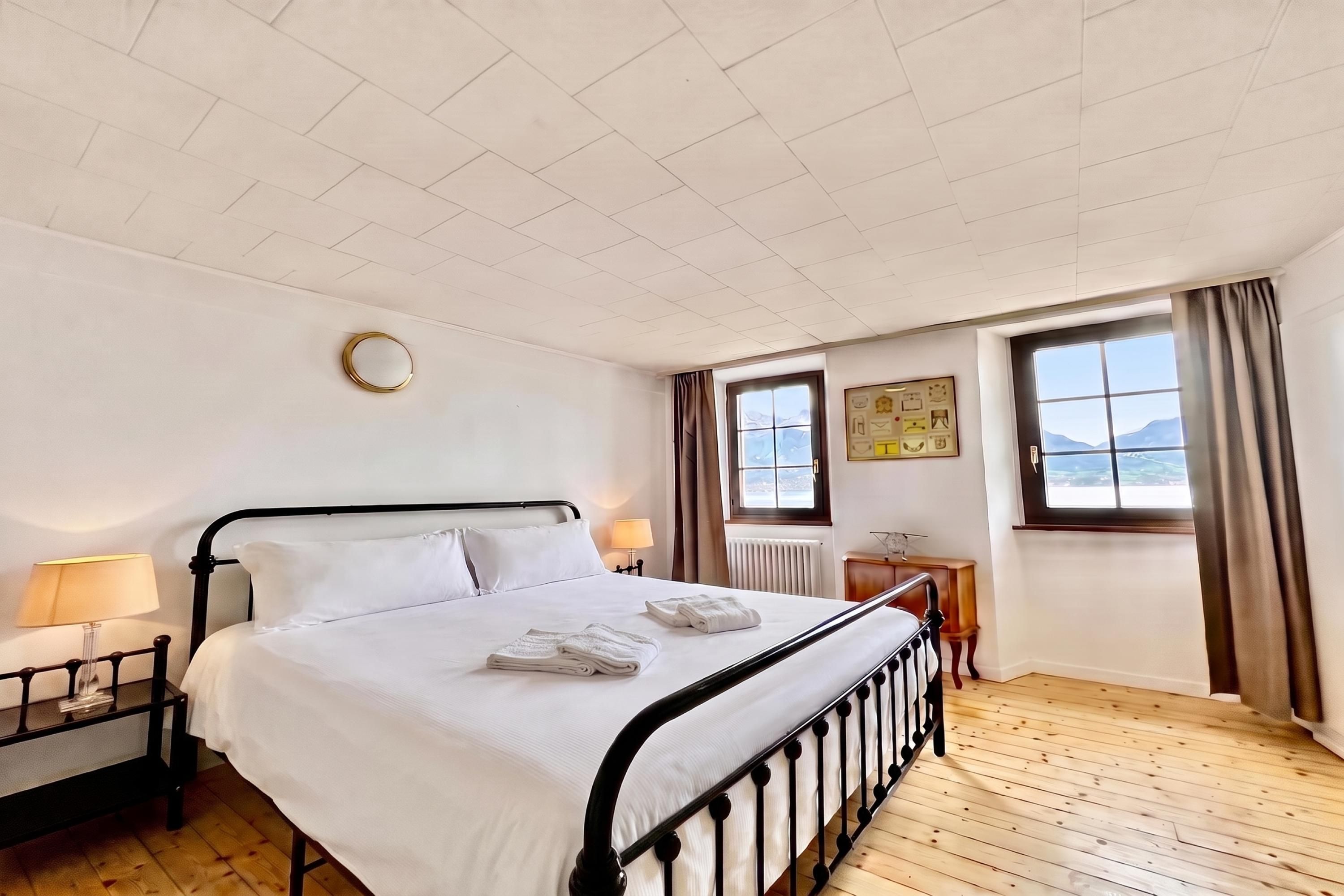 A hotel room with a bed, a bedside table with a lamp, a window with curtains, and a view of the outdoors.