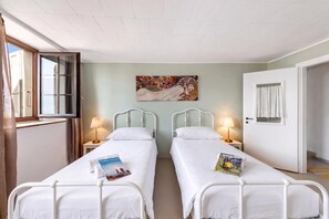 A room with two single beds, a painting on the wall, a window with curtains, and a door.