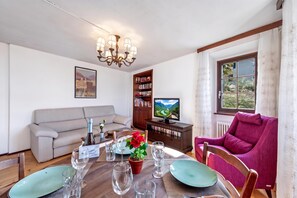 A living room with a sofa, a dining table set for two, a bookshelf, a television, and a window with curtains.