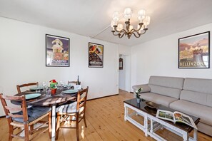A dining area with a wooden table set for four, surrounded by chairs. There is a chandelier above the table and a sofa in the adjacent living area.