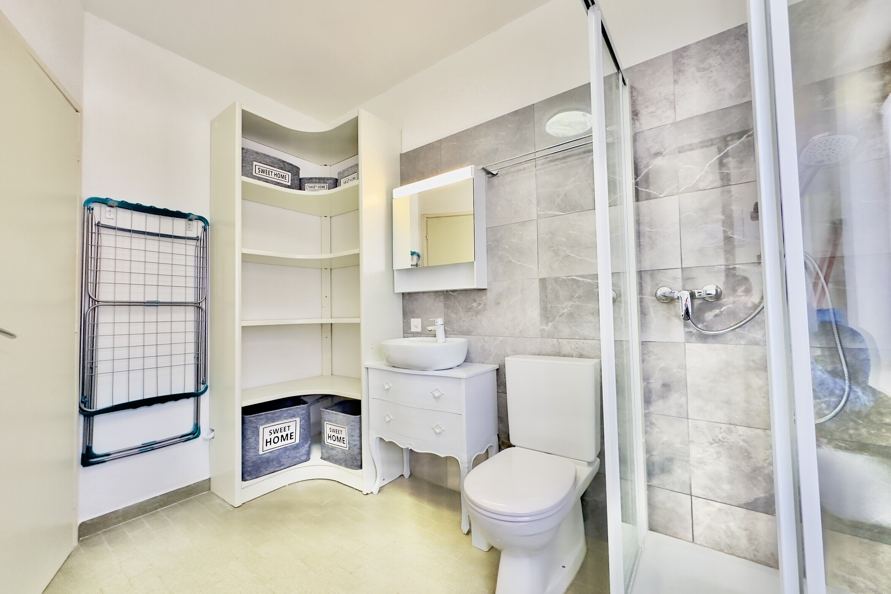 A bathroom with a toilet, sink, and shower. There are shelves and a trash bin.