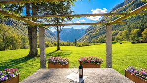 A stone table with a wine bottle and glasses, surrounded by flower boxes, under a wooden pergola with a mountain view. - Casa Mima 1 Gerra Verzasca for 4, Gerra Verzasca, Switzerland (Gerra Verzasca)