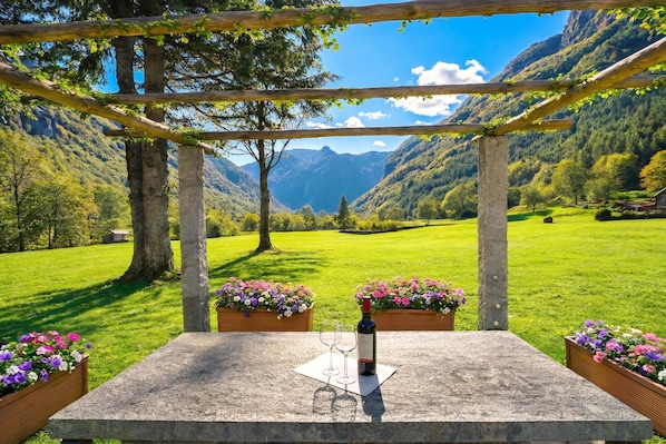 Ein Steintisch mit einer Weinflasche und Gläsern, umgeben von Blumenkästen, unter einer Holzpergola mit Blick auf die Berge.