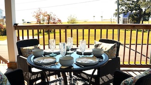A round table set for four with plates, bowls, and cutlery, surrounded by wicker chairs with patterned cushions, on a wooden deck with a view of a grassy yard and a house in the background.