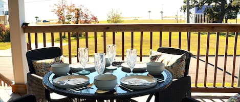 A round table set for four with plates, bowls, and cutlery, surrounded by wicker chairs with patterned cushions, on a wooden deck with a view of a grassy yard and a house in the background.