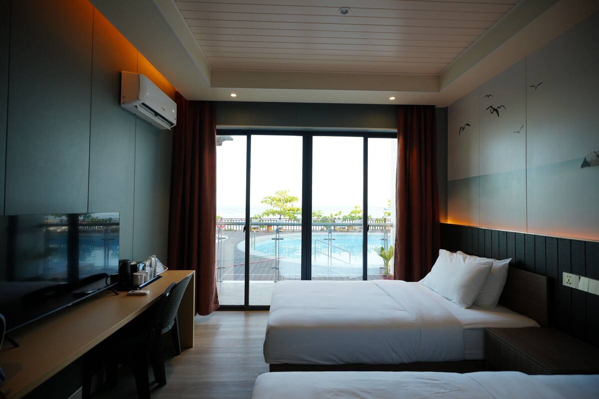 A hotel room with a bed, a desk with a TV, and a view of a swimming pool.