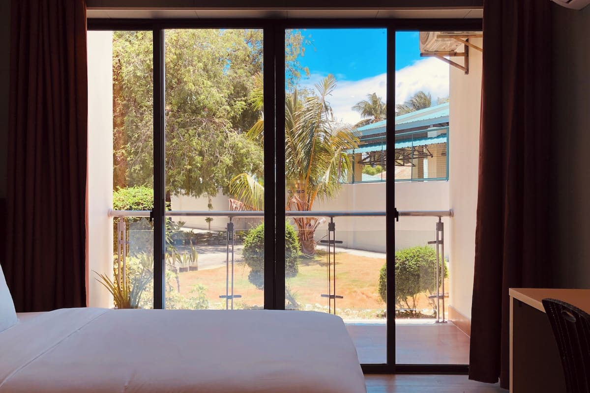 A hotel room with a large bed, a balcony with a view of greenery and a building, and a desk with a chair.