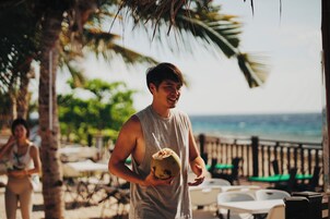Una persona sostiene una bebida de coco en una playa con palmeras y vistas al océano.