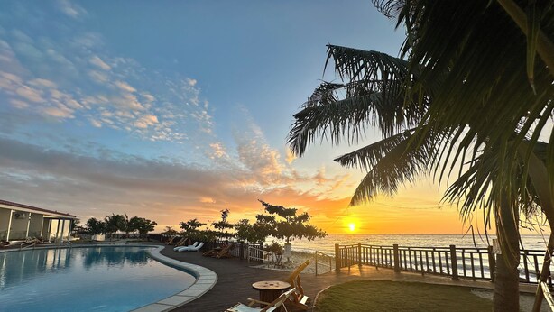 Zona de piscina con vistas a la puesta de sol sobre el océano, rodeada de palmeras y sillones de descanso.