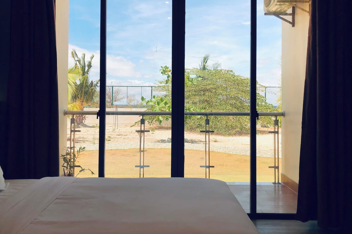 A hotel room with a large window, a bed, and a view of a fenced outdoor area with greenery.