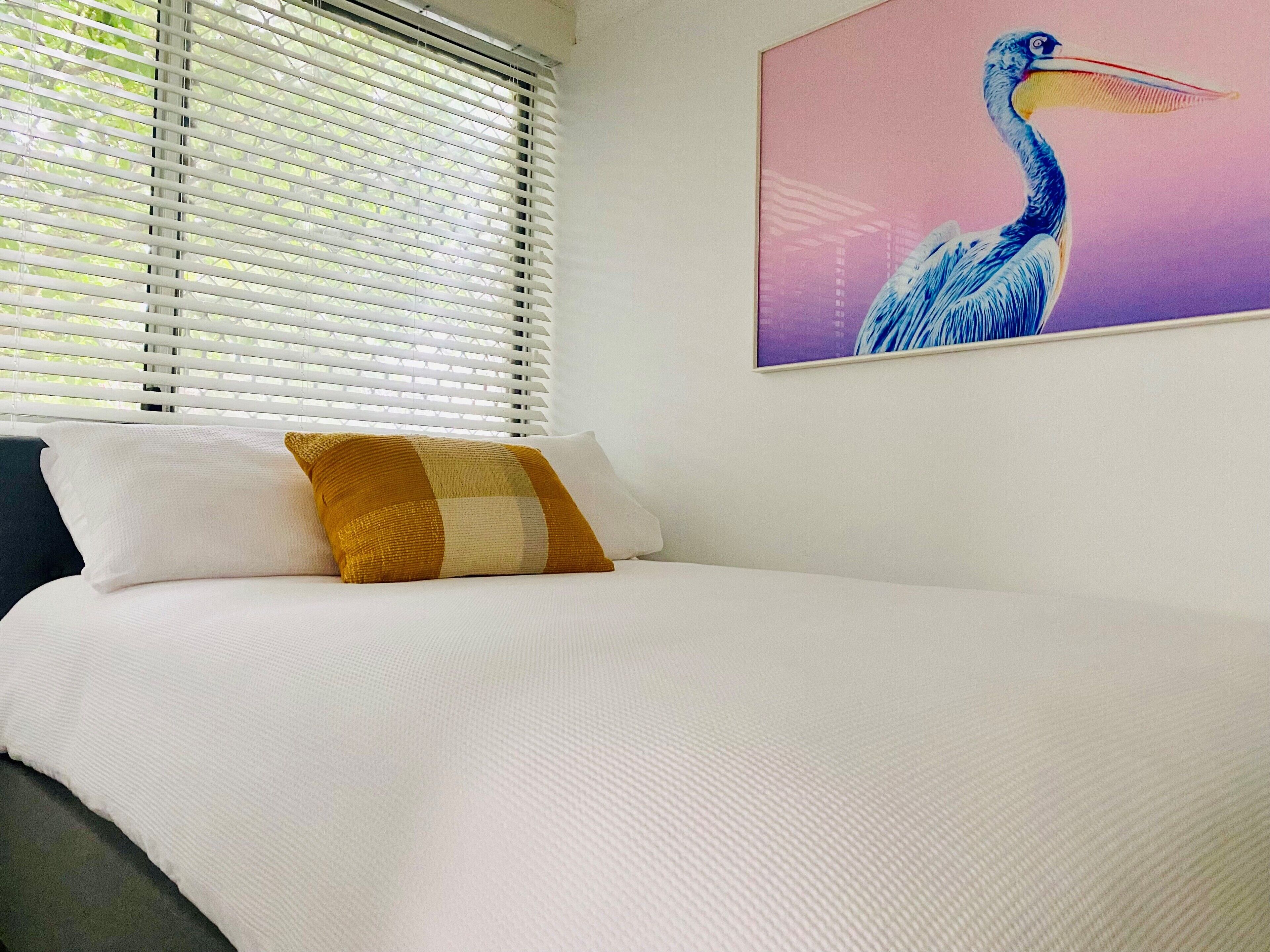 A bedroom with a bed, white bedding, a pillow with a checkered pattern, a window with blinds, and a wall-mounted painting of a bird.