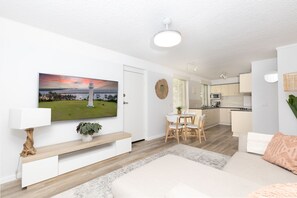 Een moderne woonkamer met een flatscreen televisie, een eethoek met tafel en stoelen, een keuken met kasten en apparatuur, en een groot raam met zonwering.