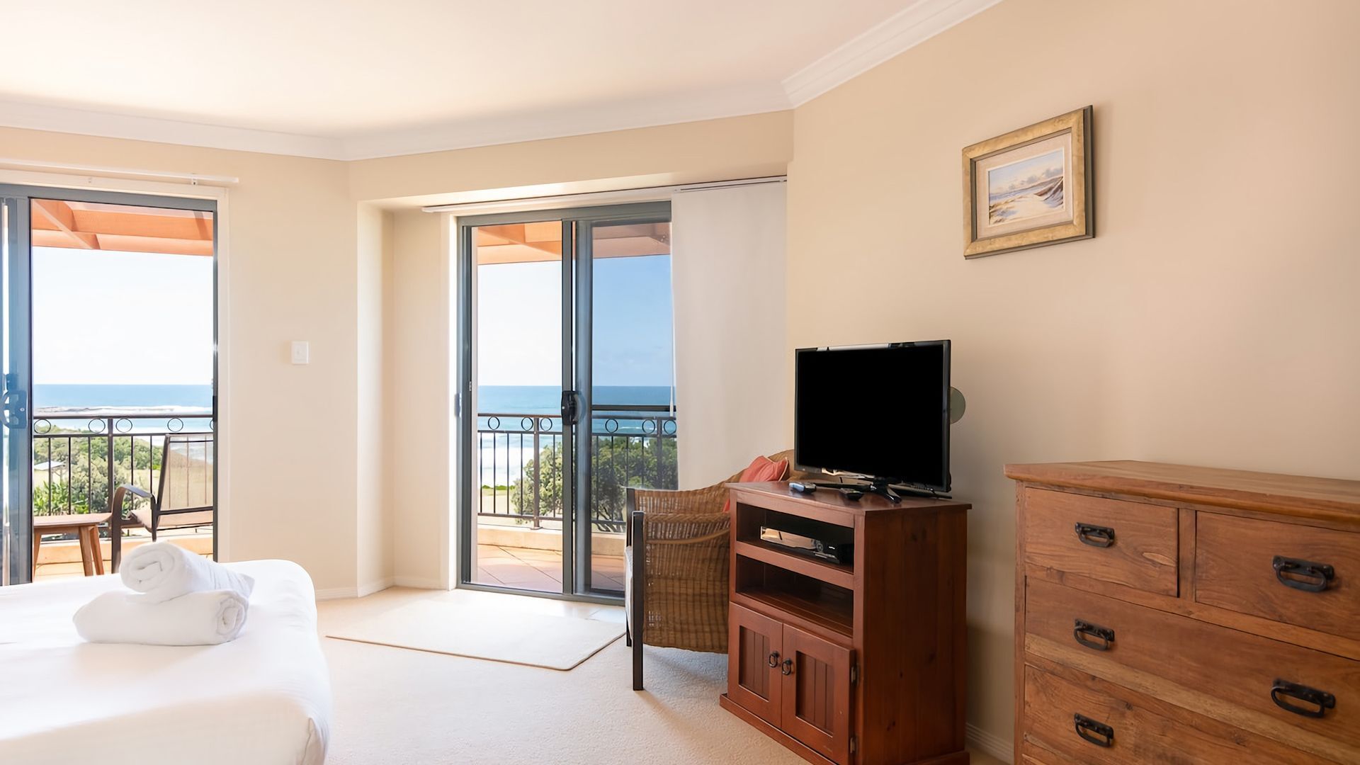 A hotel room with a bed, a television on a wooden cabinet, a chair, a balcony with a view of the sea, and a painting on the wall.