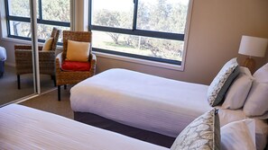A hotel room with two beds, a wicker chair, and a view of trees through large windows.
