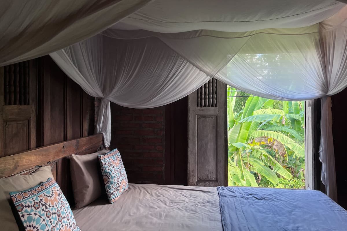 A bedroom with a bed, a wooden headboard, and a view of lush greenery through a window.