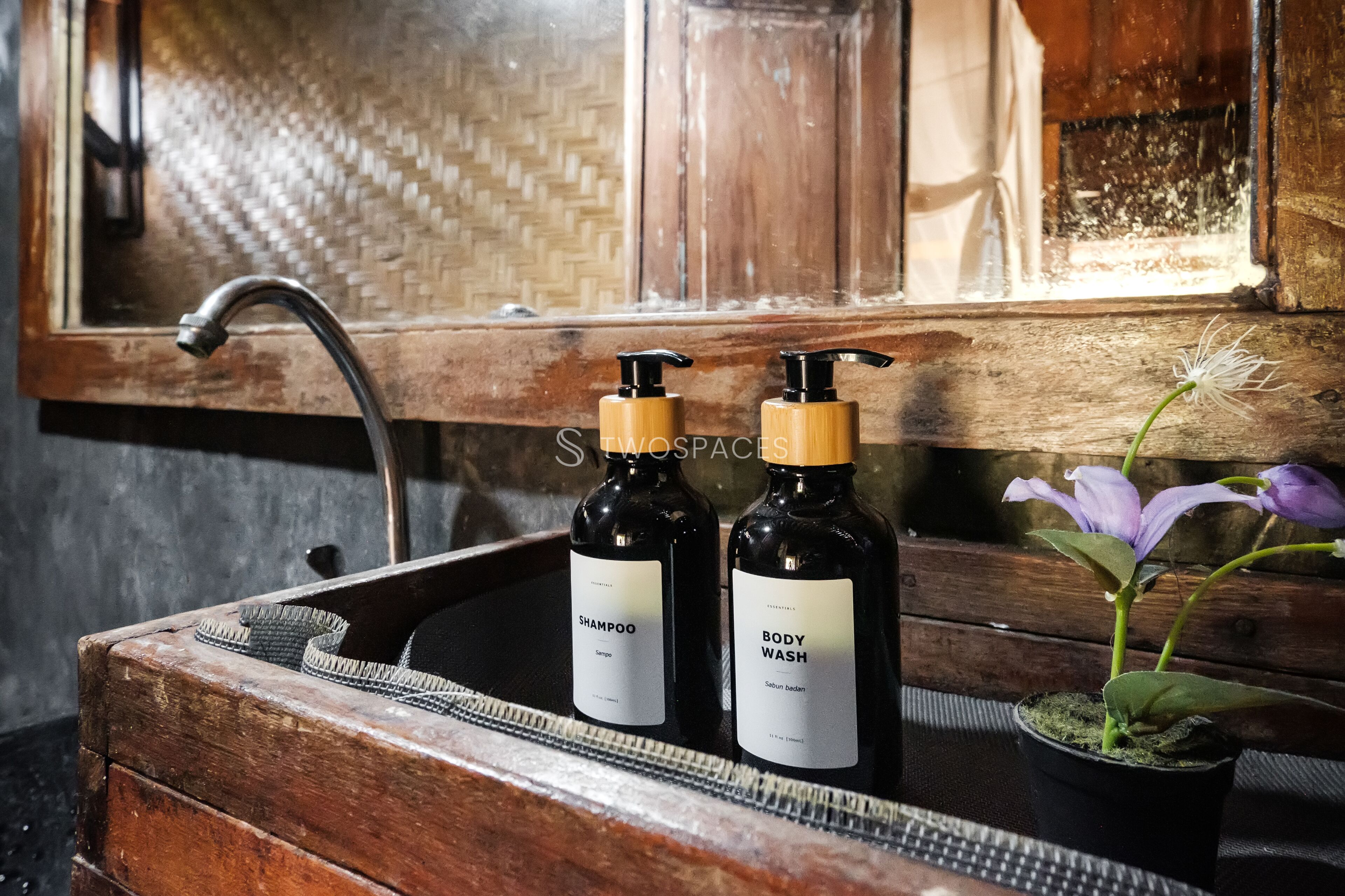 Un lavabo de madera con dos botellas de champú y gel de baño, una planta en maceta y una ventana con textura tejida.