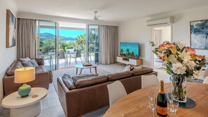 A modern living room with a sofa, a coffee table, a TV, and a dining table with wine glasses and a bottle. - Lagoon 004 by HamoRent on Hamilton Island (Hamilton Island)