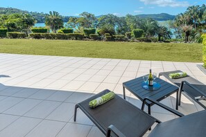Une terrasse extérieure avec un sol carrelé, une table avec une bouteille et des verres, et des chaises avec des coussins.
