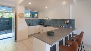 A modern kitchen with white cabinets, a large island with bar stools, a built-in oven, and a window overlooking greenery. - Lagoon 004 by HamoRent on Hamilton Island (Hamilton Island)