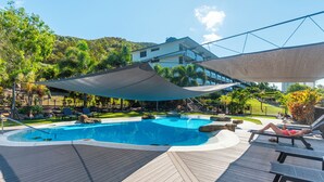 A swimming pool with lounge chairs and a shaded area, surrounded by greenery and a building in the background. - Lagoon 004 by HamoRent on Hamilton Island (Hamilton Island)