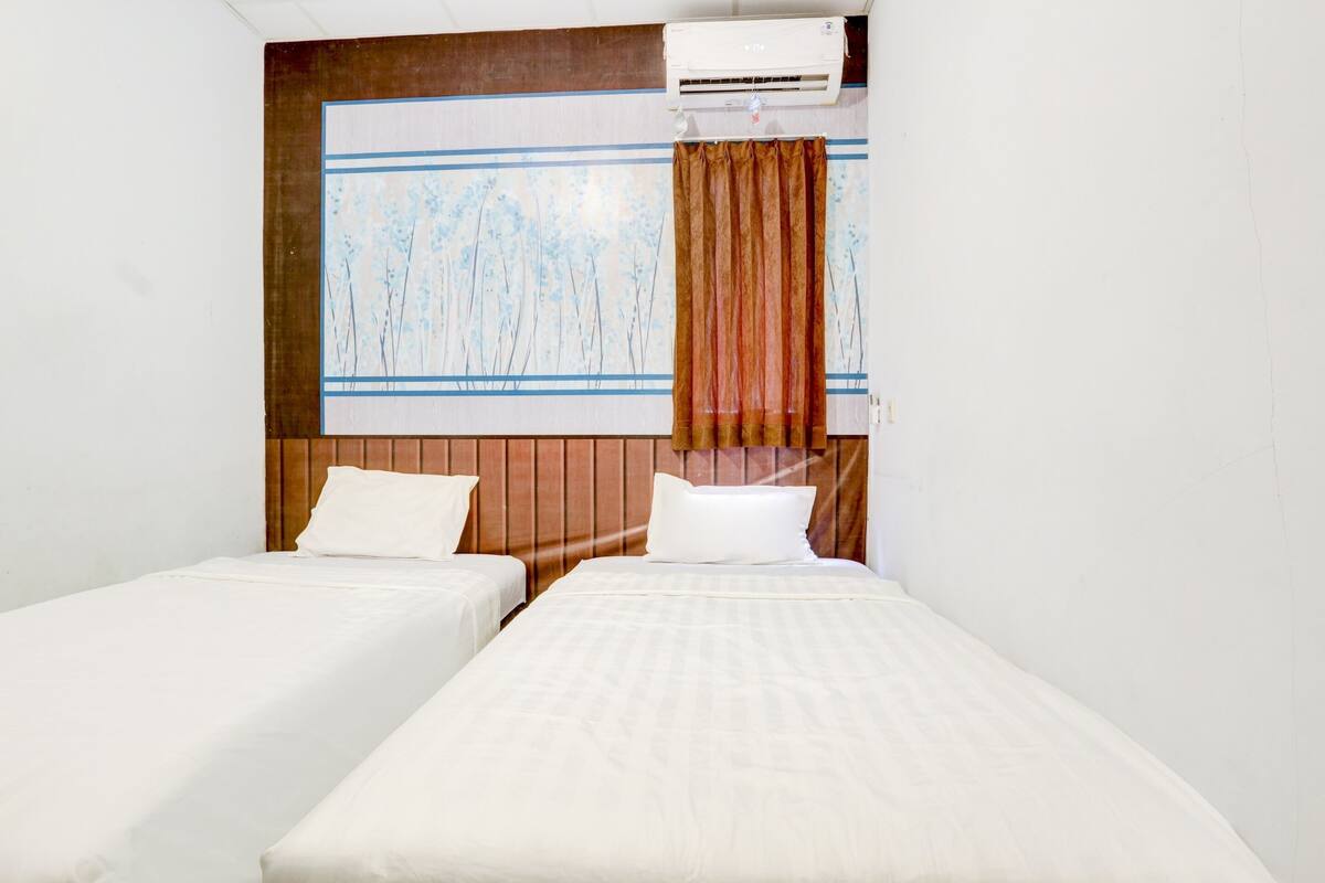 A hotel room with two single beds, a wooden headboard, a window with a view of trees, and an air conditioning unit above the window.