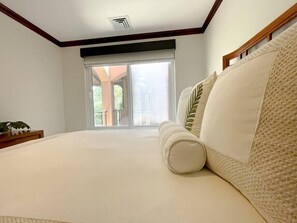 A bedroom with a large bed, a nightstand with a plant, and a view of a balcony through a sliding door.