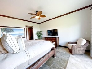 Un dormitorio con cama, ventilador de techo, televisor, una silla y un cuadro de una playa.