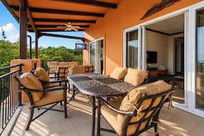 An outdoor patio with wicker furniture and a table, a ceiling fan, and a view of greenery.