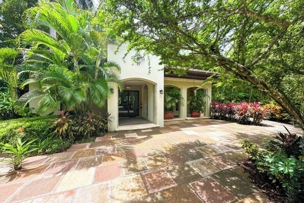 A building with a covered entrance, surrounded by lush greenery and a stone-paved pathway.