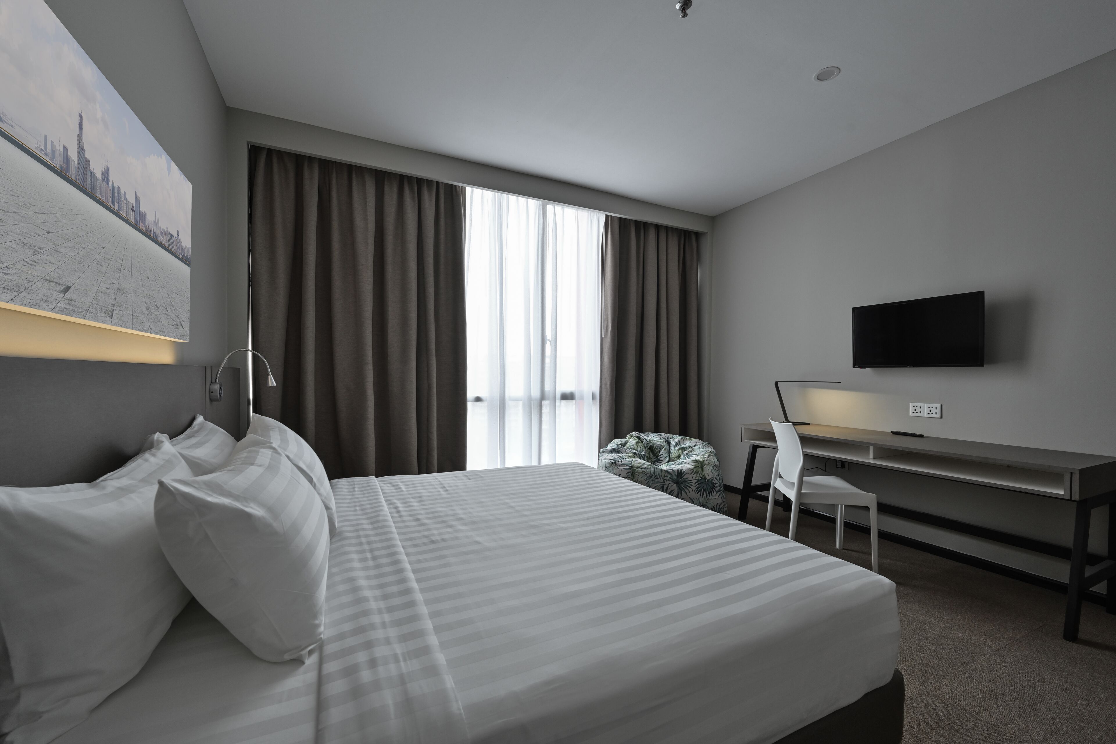 Una camera d'albergo con un letto, una scrivania, una sedia, una TV e una finestra con tende.