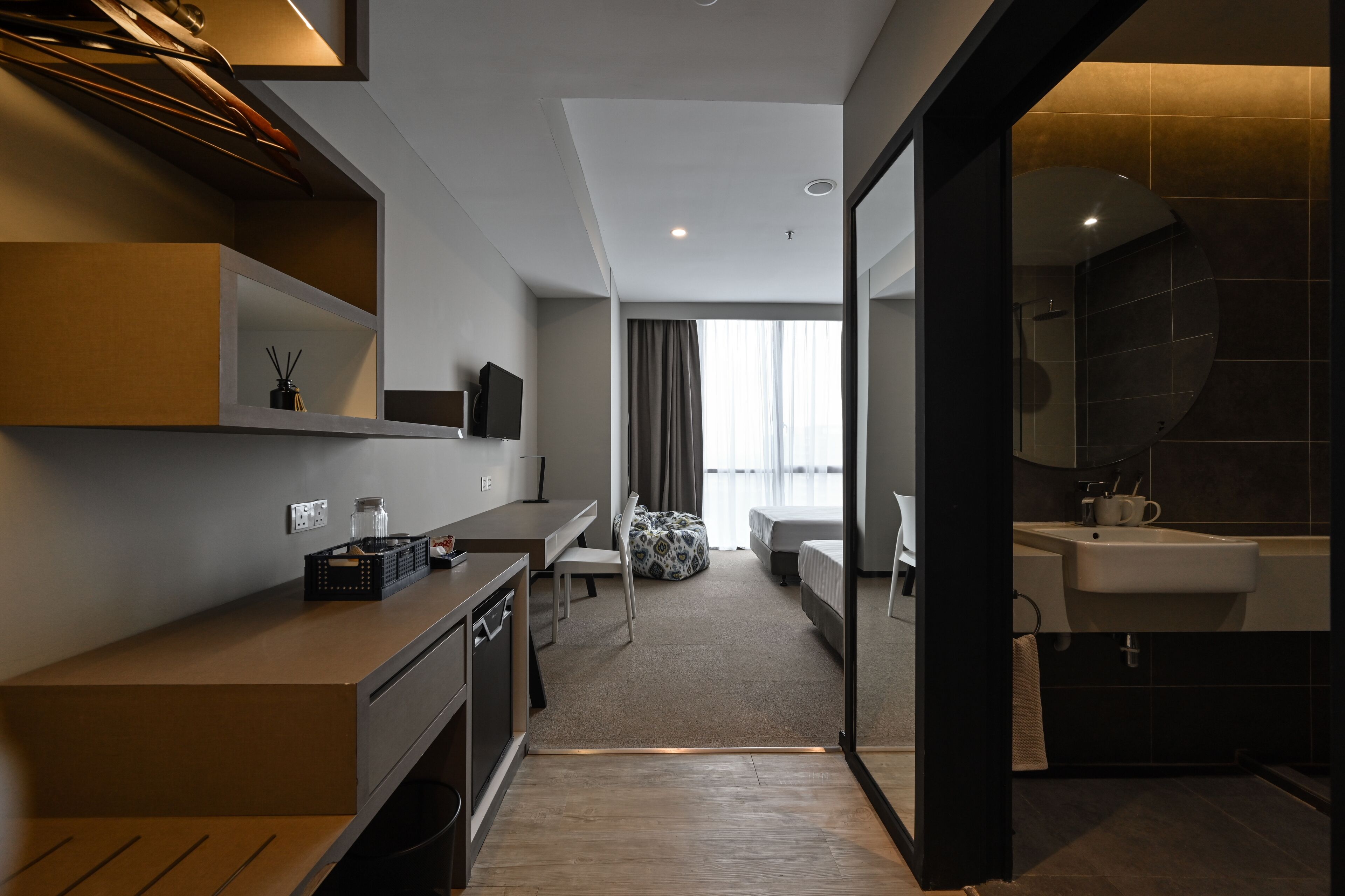 A modern hotel room with a kitchenette, a bed, a bathroom with a sink, and a large window with curtains.