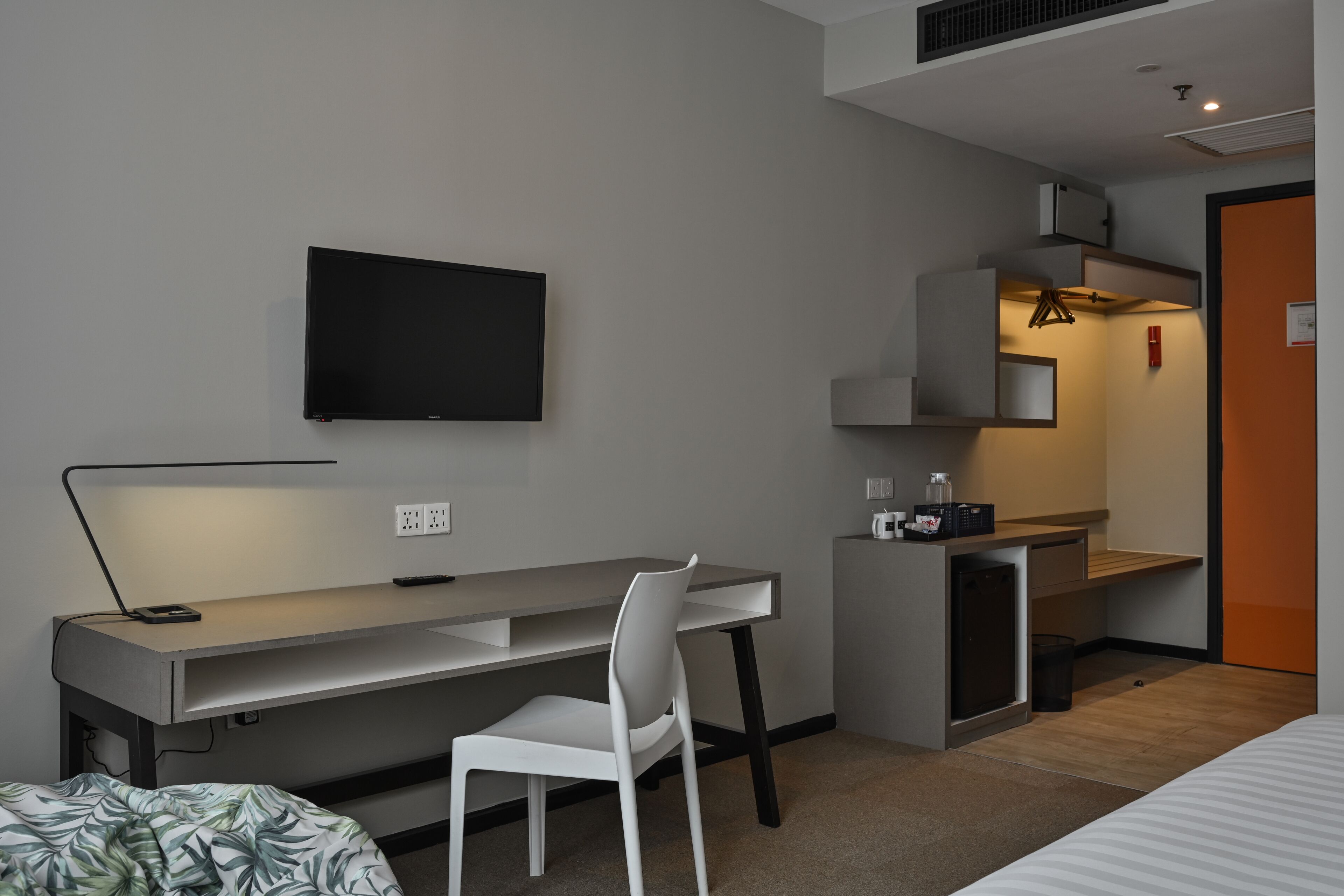 A compact hotel room with a bed, a desk with a chair, a TV mounted on the wall, and a small kitchenette area.